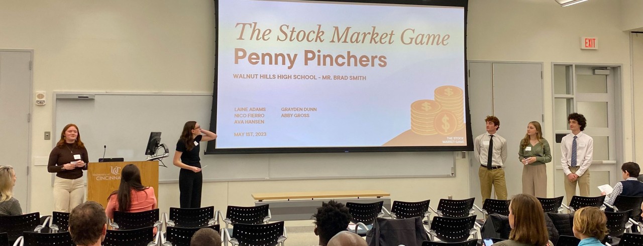 young people stand in front of a room by a screen that says the stock market game penny pinchers