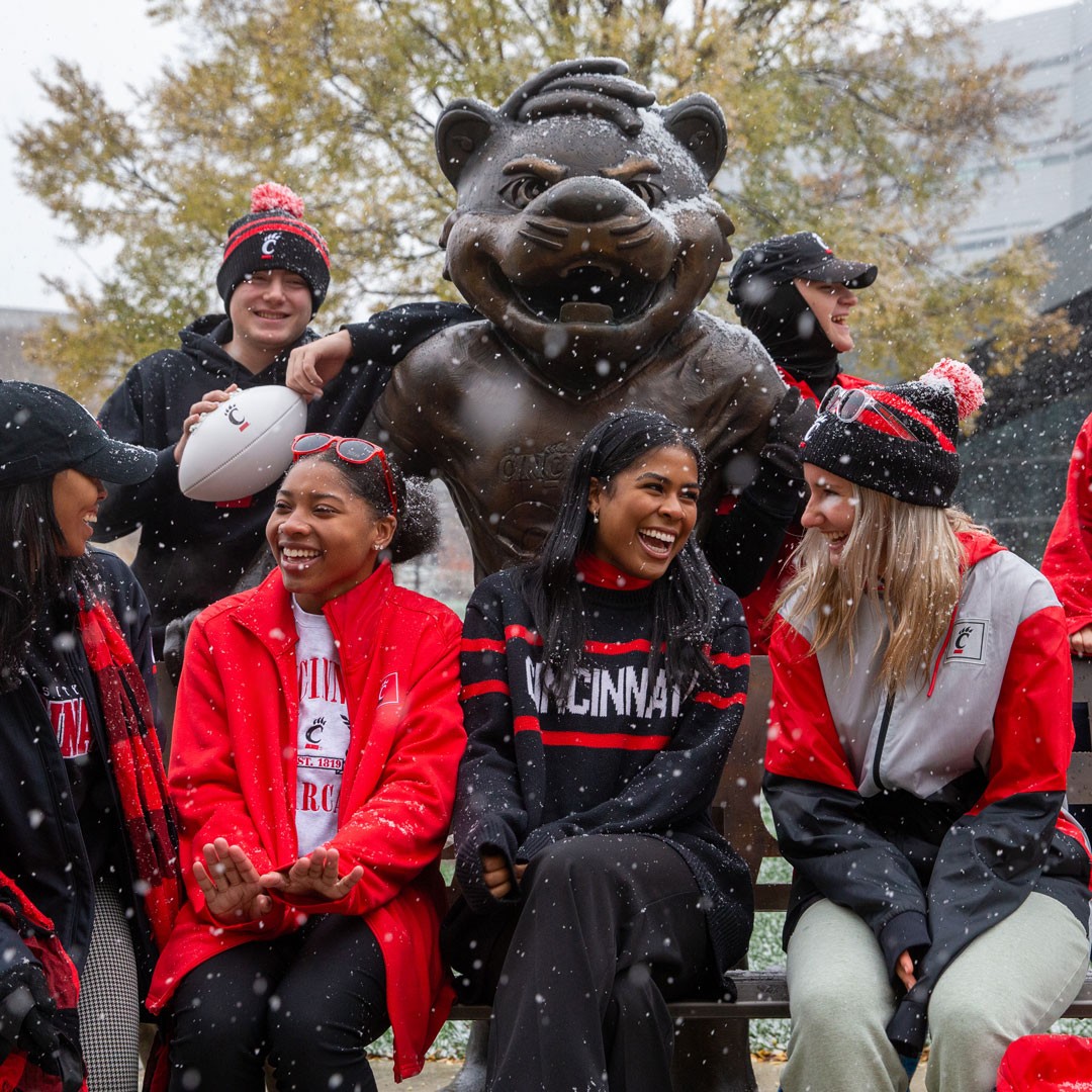 smiling students sit on a bench in front of a bearcat statue in the snow