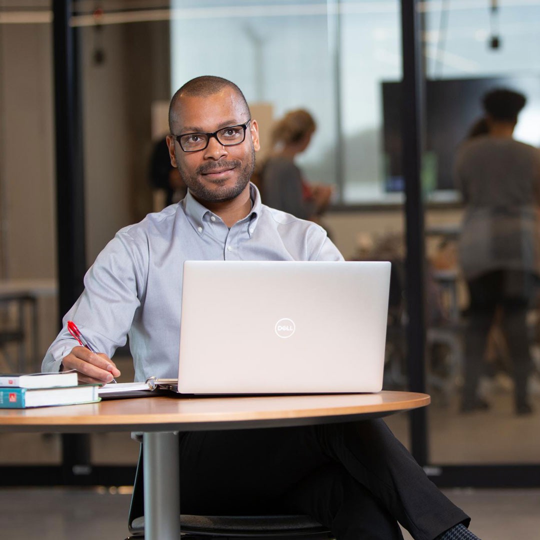 man in business clothing sits with laptop, books and pen